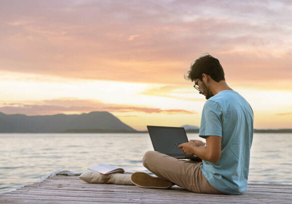 young digital nomad man sitting on wooden pier at sea working on internet remotely at sunset - Traveling with a computer - Online dream job concept