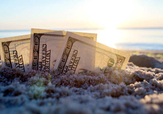 Three dollar bills are buried in sand on sandy beach near sea at sunset dawn in summer close-up. Dollar bills partially buried in sand