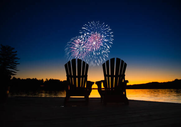 Canada day fireworks on a lake in Muskoka, Ontario Canada. On the wooden dock two Adirondack chairs are facing the sunset orange hues