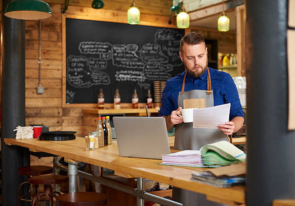 a coffee shop owner checks the delivery notes in his bookkeeping folder on the counter of his busy coffee shop and cross checks them with his online accountancy via his laptop at the end of a busy day .